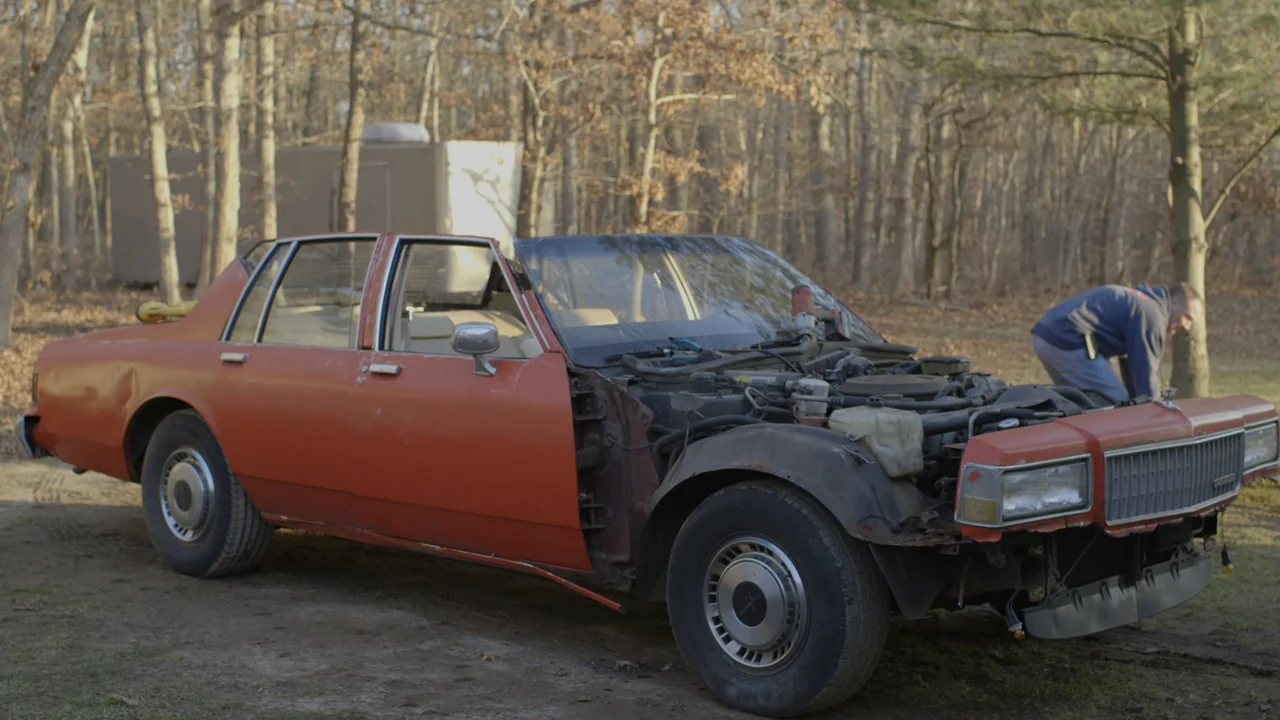 The Last Race (2022) movie still: medium — An open wooded area features a disassembled red sedan parked on dirt ground. A man leans over the exposed…