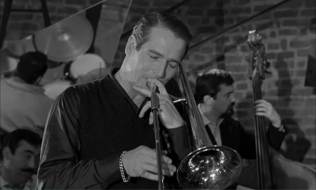 Paris Blues (1961) movie still: medium — A dimly lit room with exposed brick walls and a jazz band performing. The main subject in a dark vest plays…