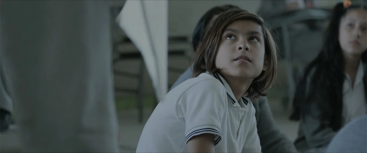 Radical (2023) movie still: medium — A group of children sit together inside a brightly lit school classroom environment. The central child gazes…