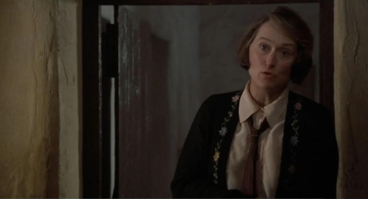Dancing at Lughnasa (1998) movie still: medium — A woman stands in a dimly lit doorway framed by textured walls. She wears a floral cardigan and…