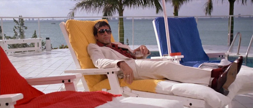 Scarface (1983) movie still: medium — A sunny pool deck overlooks a calm body of water with a distant city skyline. A man wearing a cream suit rests…