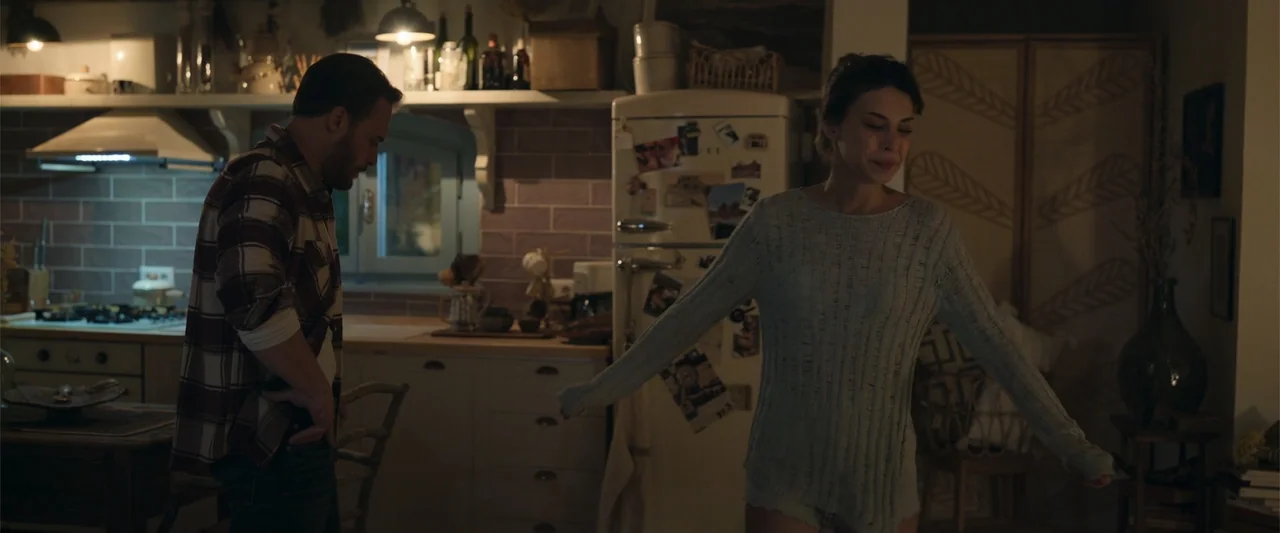 Blue Cave (2024) movie still: medium — A residential kitchen features white cabinetry and a tiled backsplash wall. A man and a woman stand together…