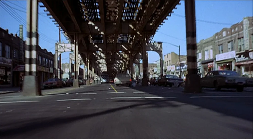 The French Connection (1971) movie still: establishing — An urban city street is framed beneath a massive metal elevated train track structure.…