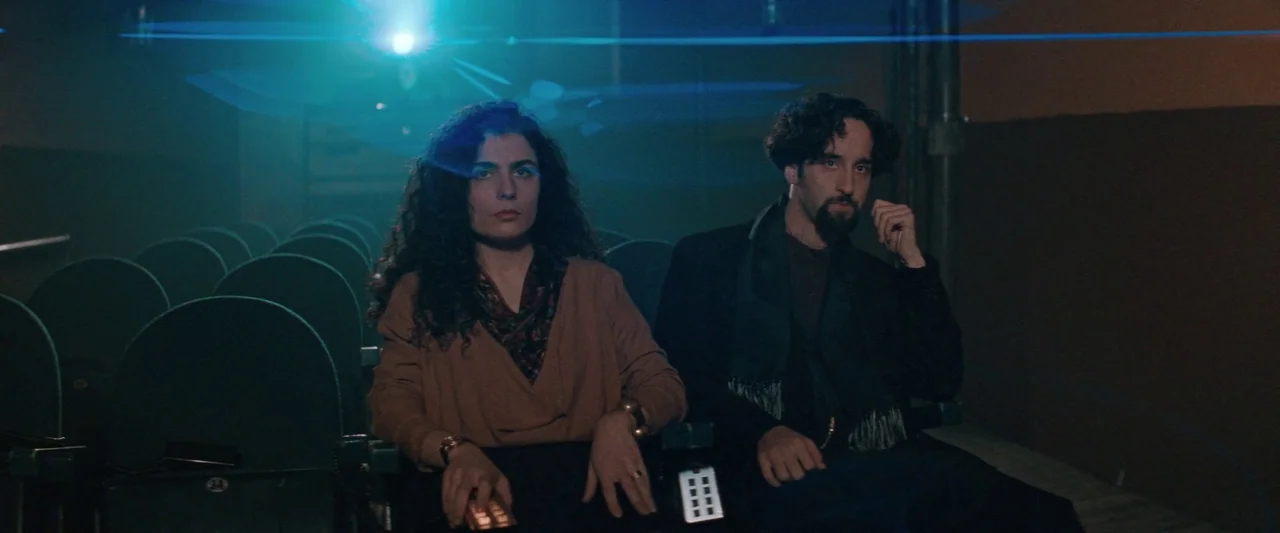 The Adjuster (1991) movie still: medium — Two individuals sit in a dark movie house surrounded by rows of empty seats. The man and woman face…