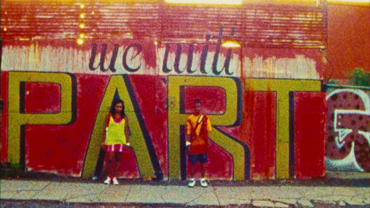 Red Hook Summer (2012) movie still: wide — A textured wall features large painted yellow letters on a vibrant red background. Two individuals stand…