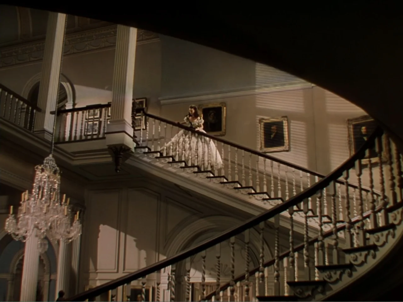 Gone with the Wind (1939) movie still: wide — A grand mansion interior features an expansive curved wooden staircase and a large chandelier. A woman…