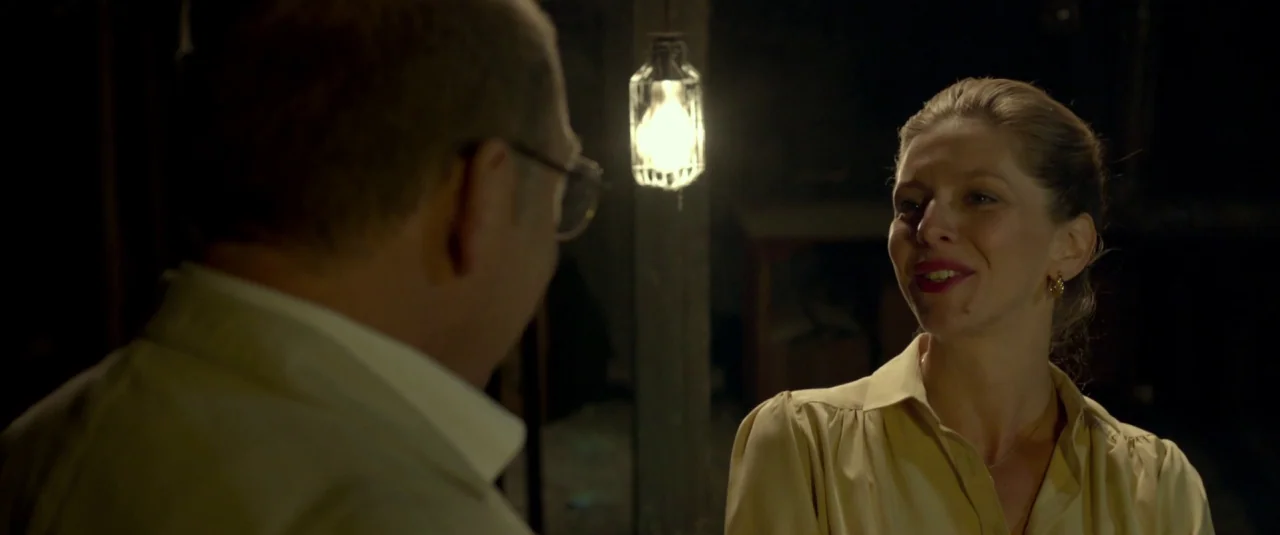 Big Bad Wolves (2013) movie still: medium — A dimly lit room contains a man and a woman in conversation. The woman faces the man while he is viewed…