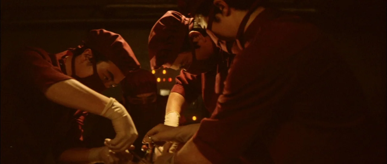 Terror of Mechagodzilla (1975) movie still: medium — Three surgeons in dark red uniforms lean over a central point in a dark room. They wear…