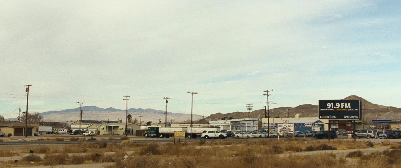 To Leslie (2022) movie still: establishing — A wide view of a dry, arid desert town with low-rise buildings and mountains. Telephone poles,…