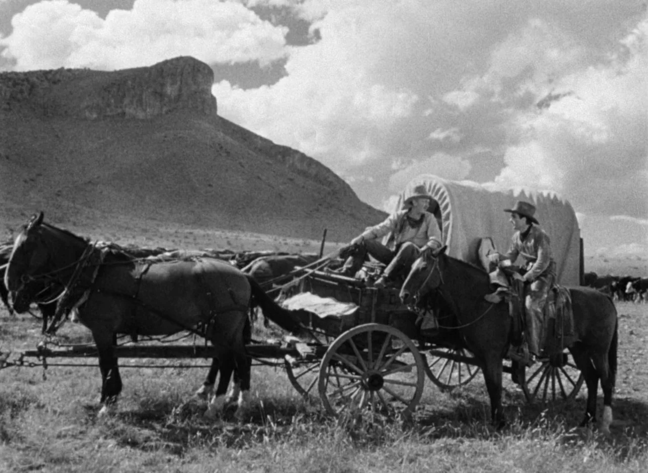 Red River (1948) movie still: wide — A dry, grassy plain sits beneath a large rocky formation and scattered white clouds. Two men with a horse-drawn…