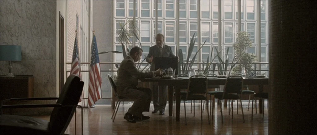 Tinker Tailor Soldier Spy (2011) movie still: wide — A modern 1970s office conference room features wood flooring and a large glass window wall. Two…