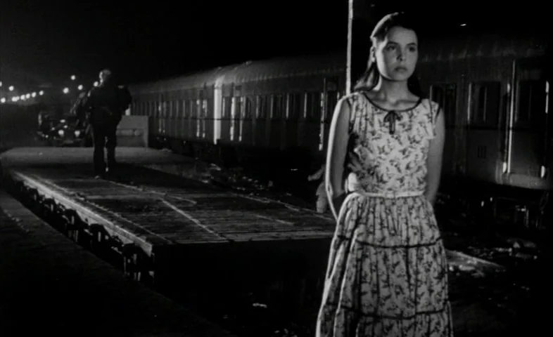 Cairo Station (1958) movie still: fullbody — A dark, empty train station platform at night with passenger carriages in the background. A woman in a…