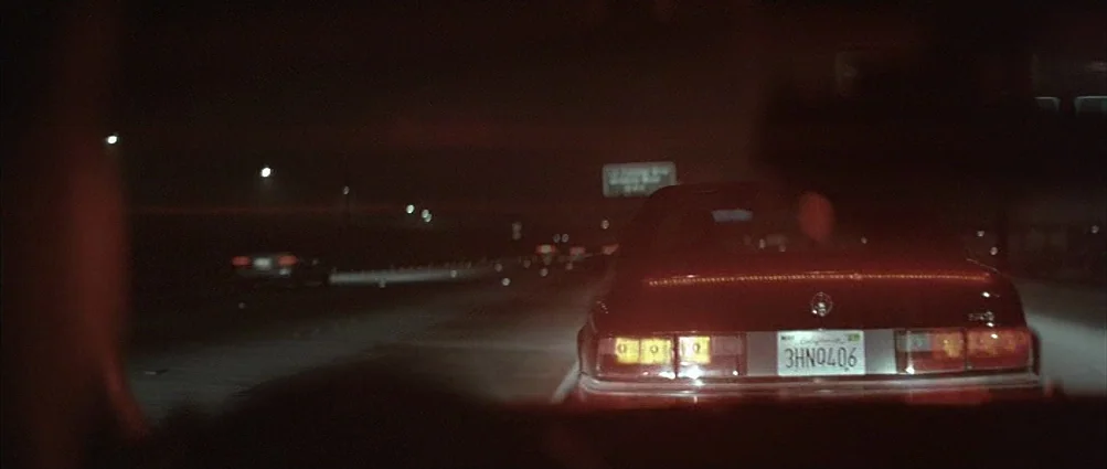 Heat (1995) movie still: wide — A dark, multi-lane highway at night is sparsely populated with vehicles. A red sedan occupies the center of the…