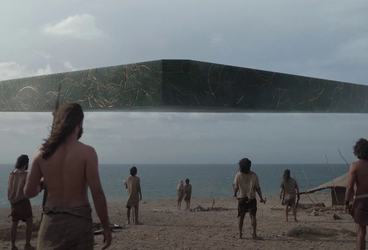 Eternals (2021) movie still: wide — A group of people wearing simple primitive clothing stand on a sandy beach. They watch a massive, dark green…