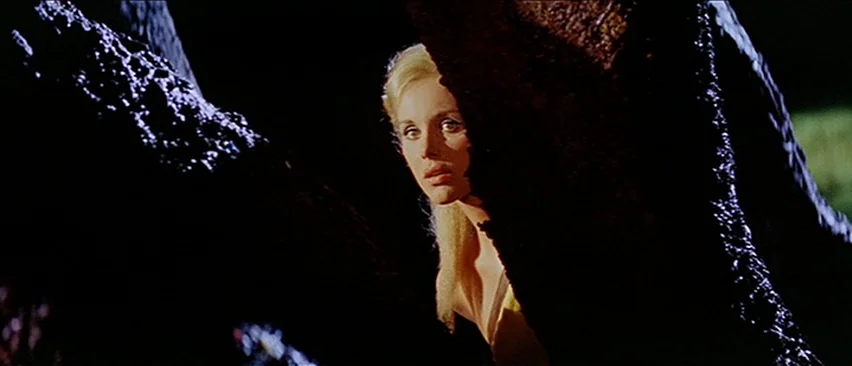 Erik The Conqueror (1961) movie still: medium — A dark, wooded area features large tree trunks framing the composition in deep shadow. A blonde…