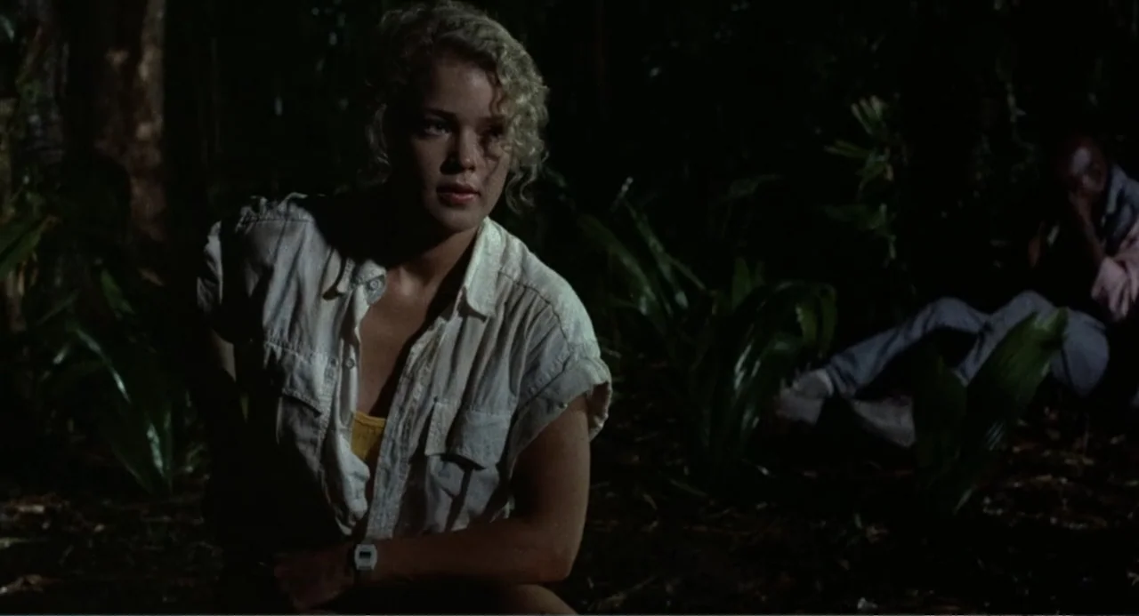 Firewalker (1986) movie still: medium — A dense, dark jungle environment at night features lush tropical foliage and thick brush. A blonde woman in…