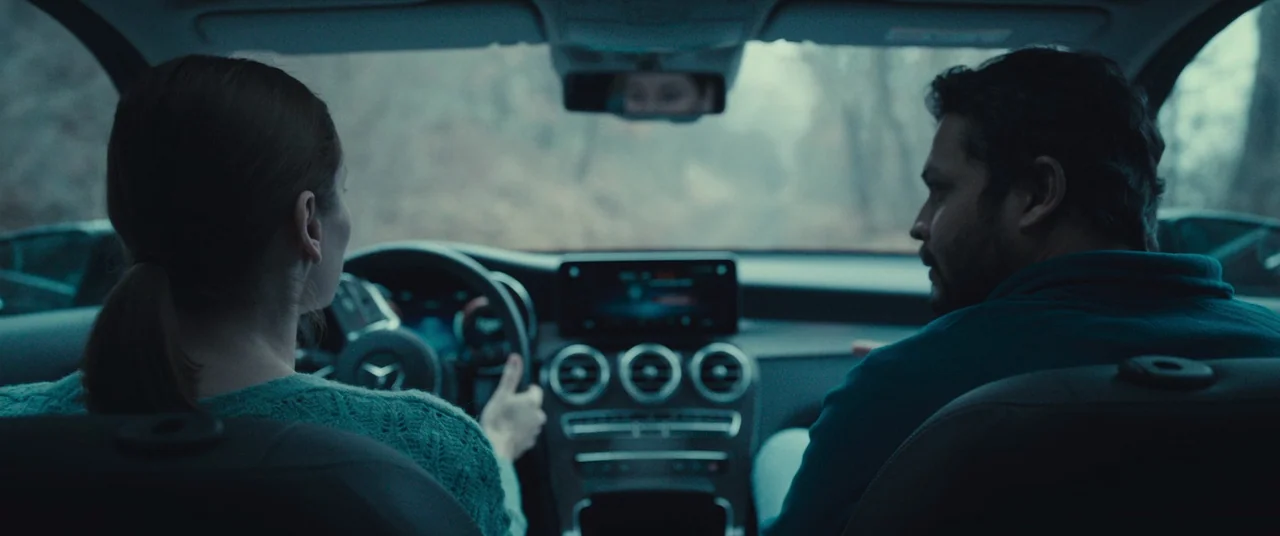 R.M.N. (2022) movie still: medium — The interior of a modern Mercedes-Benz vehicle with a view of a wooded area. A man and woman sit in the front…