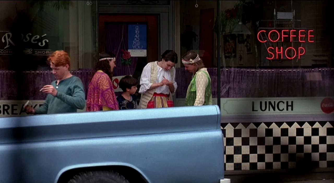 Dogfight (1991) movie still: medium — A sidewalk storefront displays a neon sign and checkered tile beneath a large glass window. A group of young…