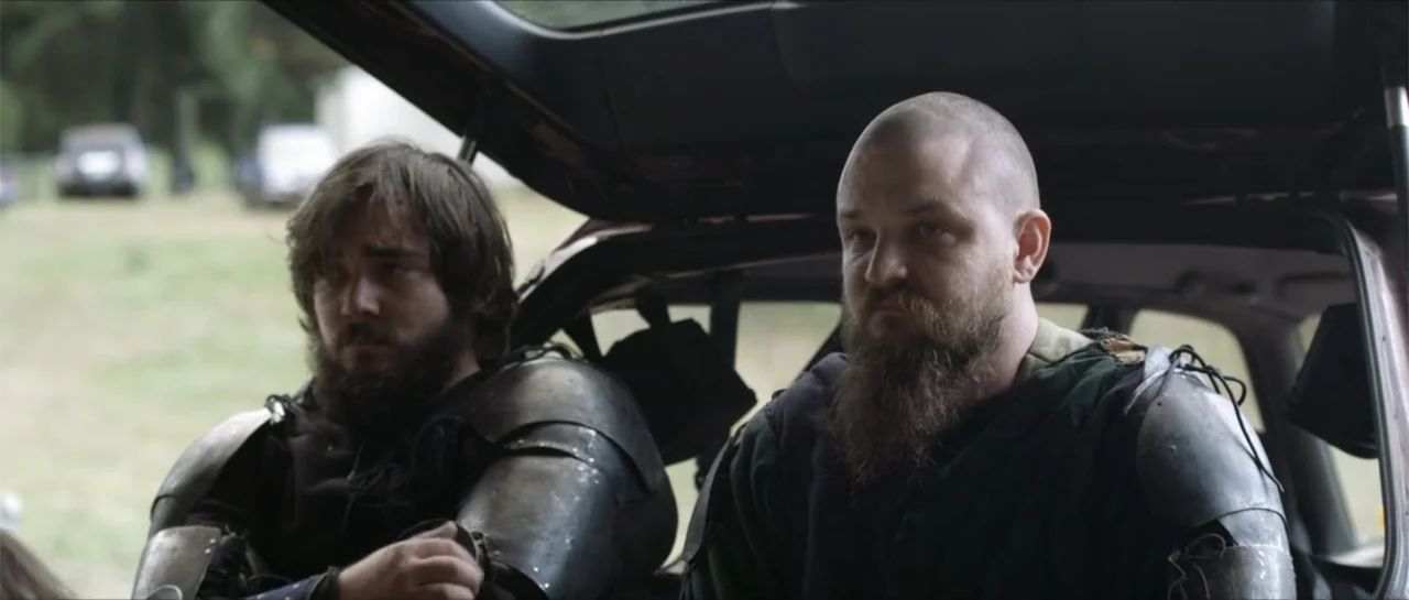 Bludgeon (2018) movie still: medium — Two men in medieval plate armor sit inside the open trunk of a hatchback. The men face forward toward the…