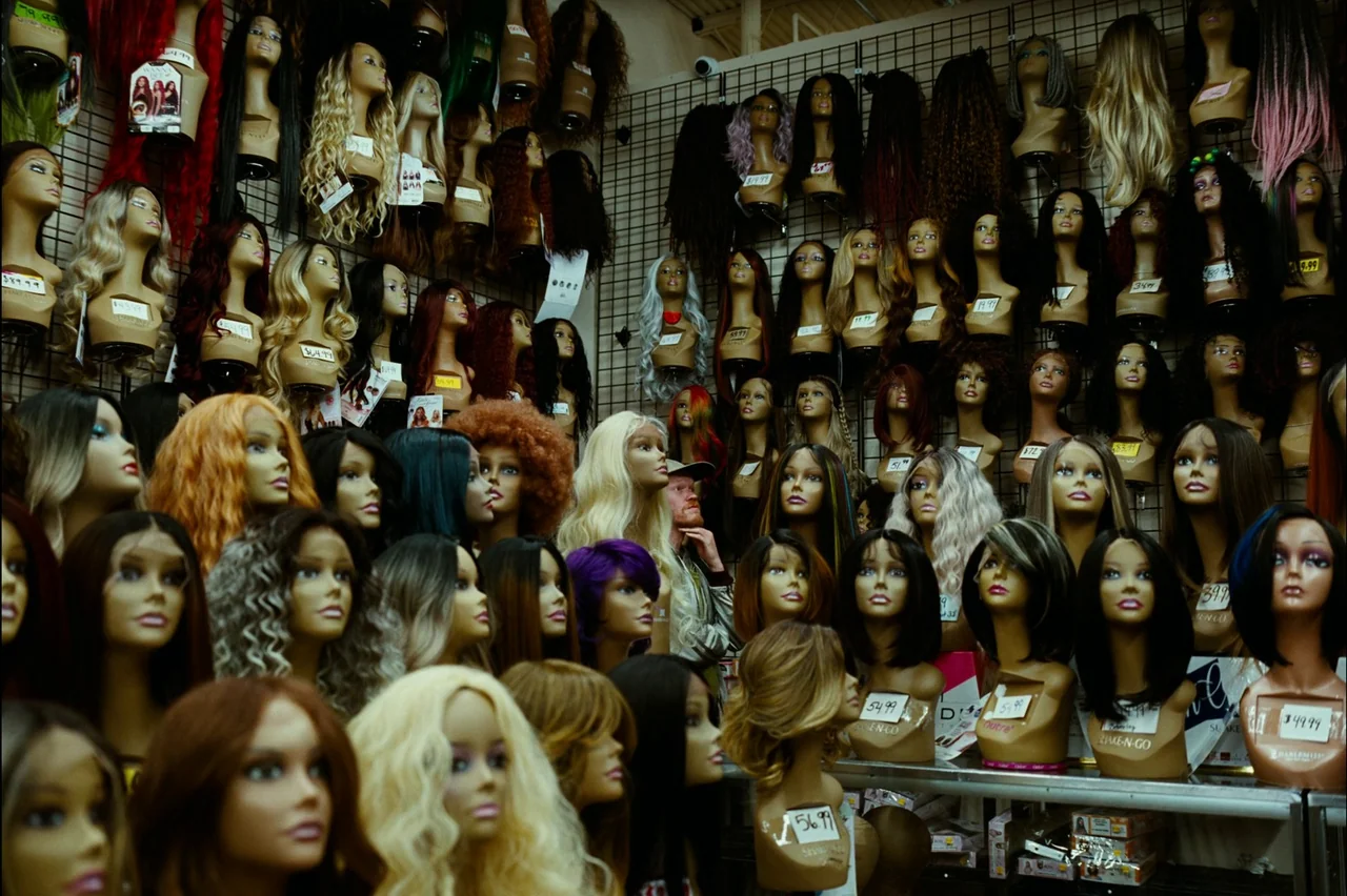 Bugonia (2025) movie still: medium — A dense collection of mannequin heads wearing various wigs occupies shelves in a store. A man stands behind the…