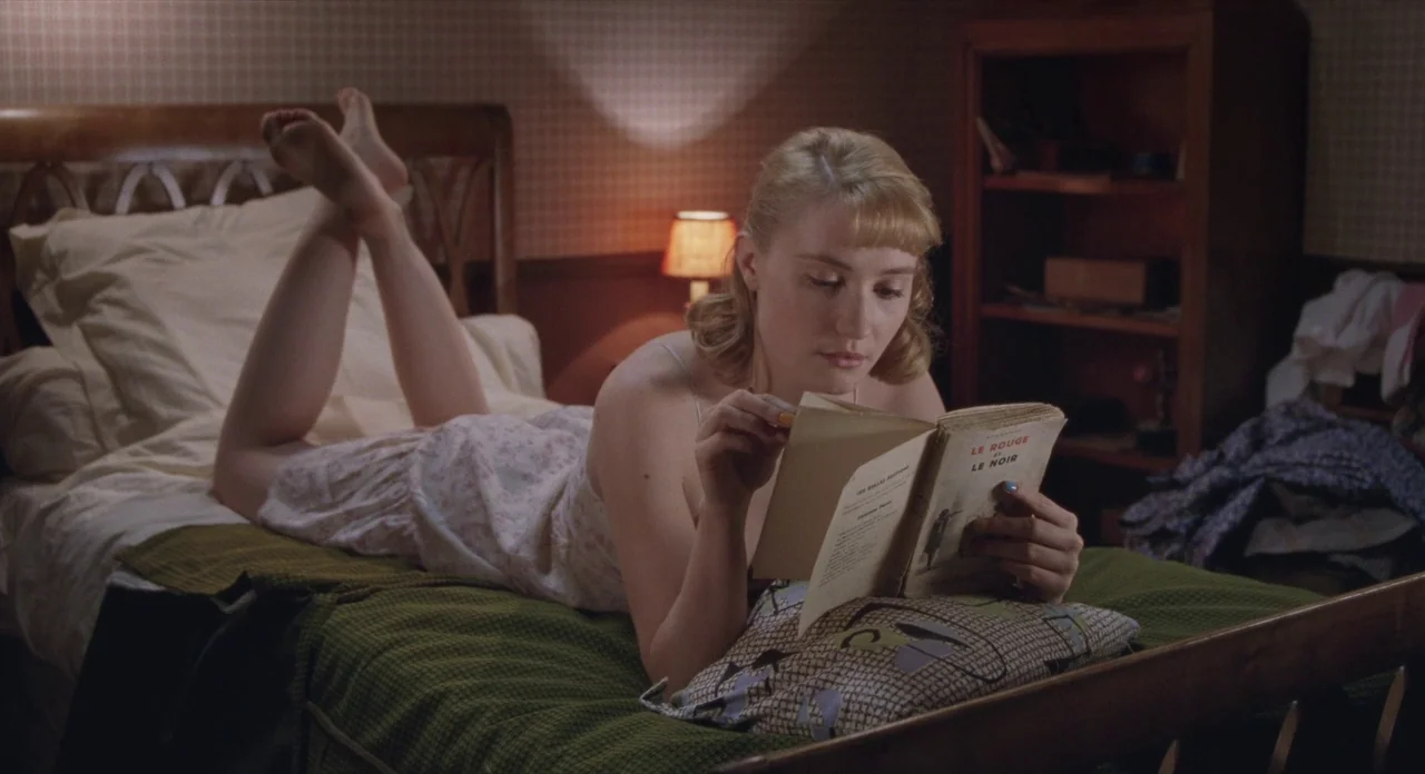 Populaire (2012) movie still: medium — A woman lies on a bed reading a book in a sparsely furnished room. The wallpaper has a subtle grid pattern…