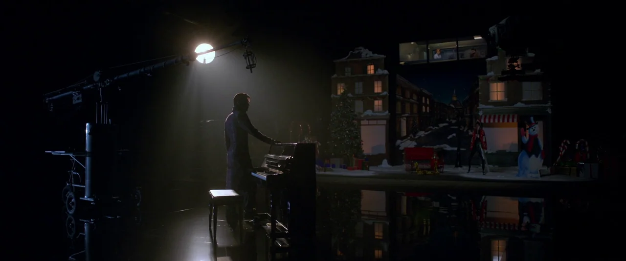 Elvis (2022) movie still: wide — A vast black sound stage features a piano and a miniature Christmas village backdrop. A man in a dark coat sits at…