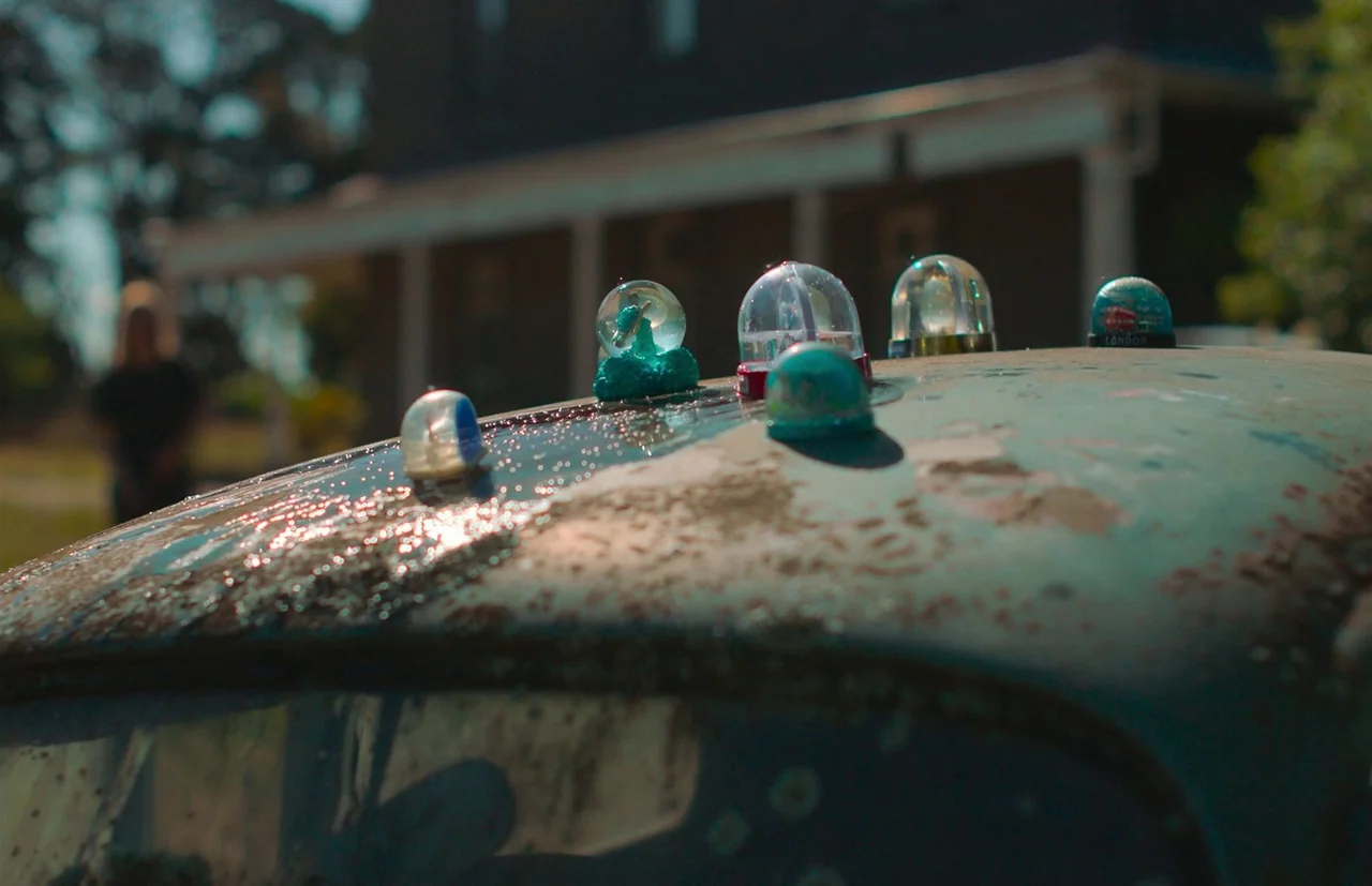 Nitram (2021) movie still: closeup — Five miniature glass snow globes are arranged on the weathered, oxidized metal of a car. The hood is covered in…