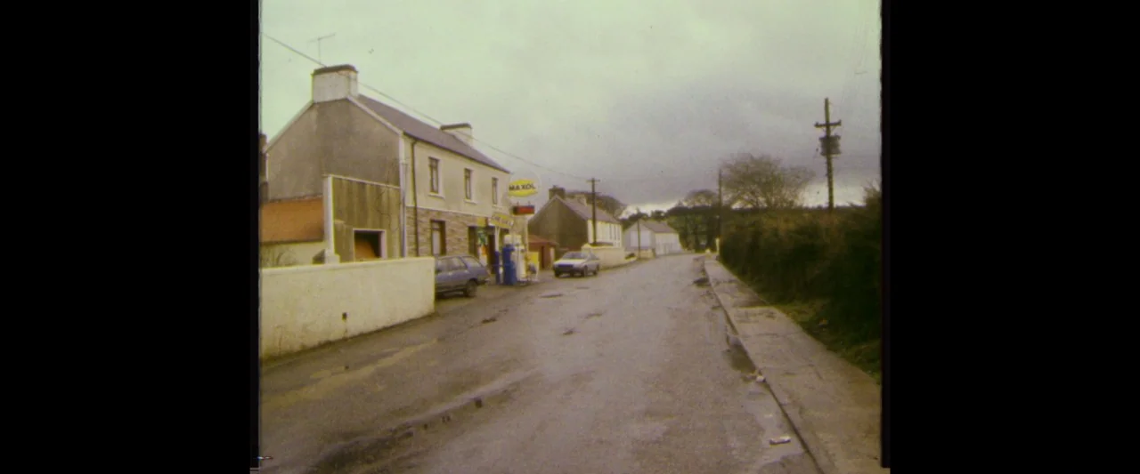 Extra Ordinary (2019) movie still: wide — An overcast, rural road in Ireland features a small Maxol gas station. A weathered stone building and a…