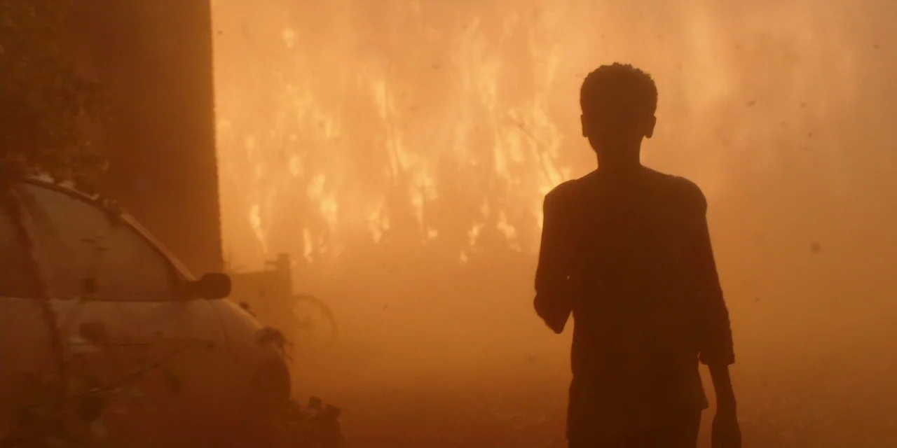 The Girl With All The Gifts (2016) movie still: medium — A figure stands in a hazy area with smoke filling the warm, firelit air. A parked vehicle…
