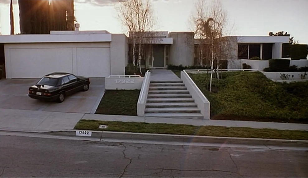 Safe movie still: establishing — A modernist one-story residence with a concrete stairway leads to the entrance. A black sedan is parked in the…