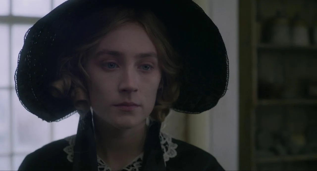 Ammonite (2020) movie still: closeup — A close portrait of a young woman in 19th-century attire inside a room. She faces forward while wearing a…
