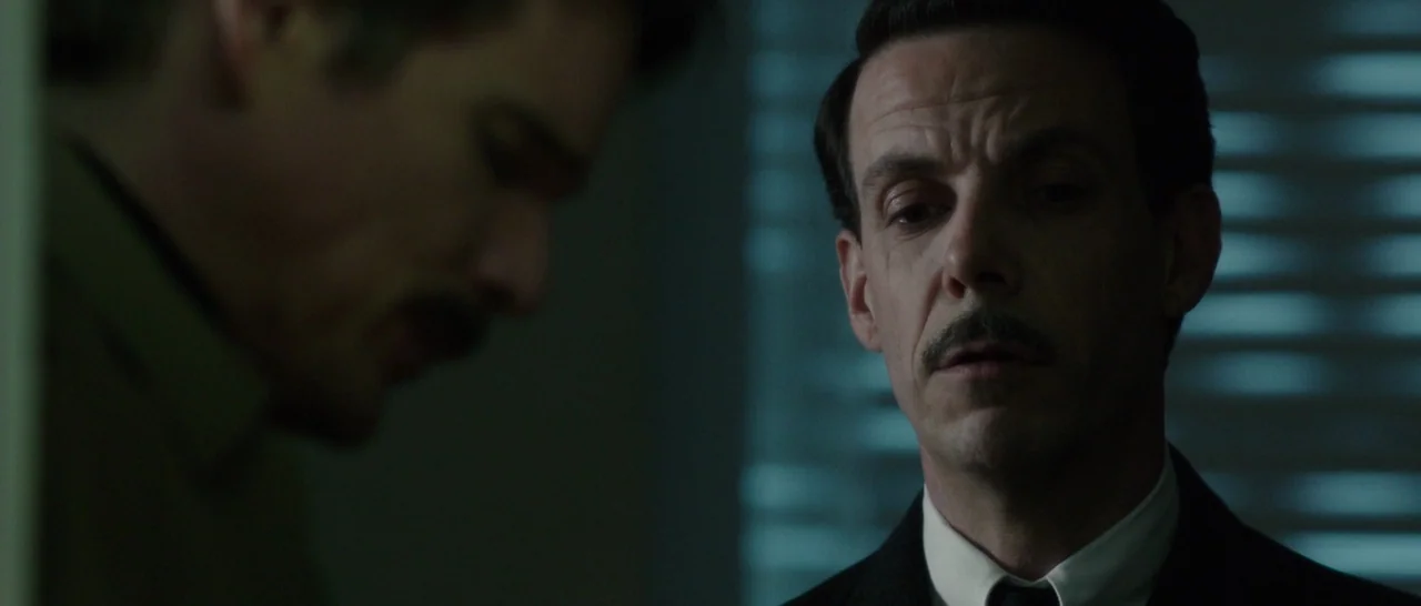 Predestination (2014) movie still: medium — A dimly lit office interior with slatted blinds visible in the background. A man in a suit faces another…
