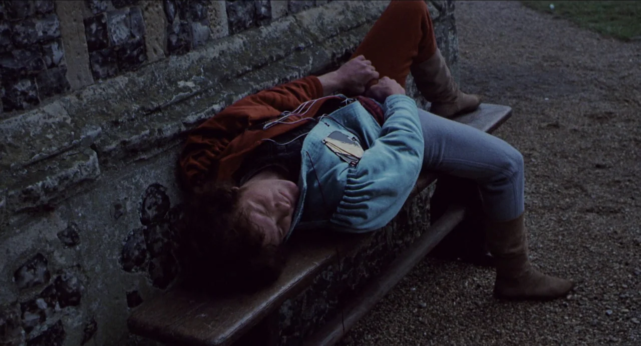 The Canterbury Tales (1972) movie still: fullbody — A person lies on a wooden bench placed against a rustic flint stone wall. The individual is…