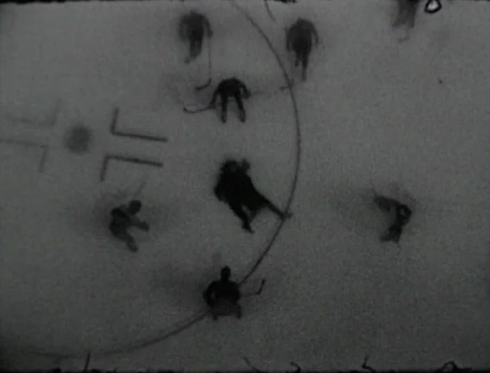 Cowards Bend The Knee (2003) movie still: establishing — An aerial view of an ice hockey rink shows players clustered at center ice. The grainy,…