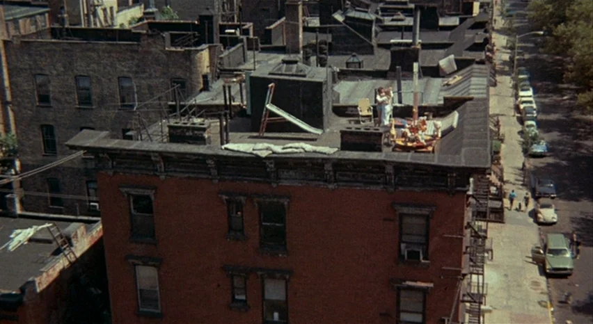A Safe Place (1971) movie still: establishing — A dense urban rooftop with a small slide and folding chairs sits above brick buildings. A narrow…