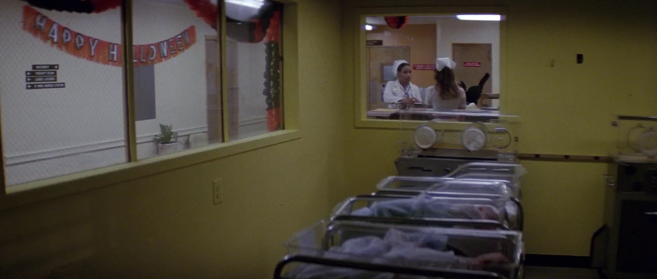 Halloween II (1981) movie still: wide — A hospital nursery features rows of plastic bassinets and yellow-painted walls. Two nurses stand in the…