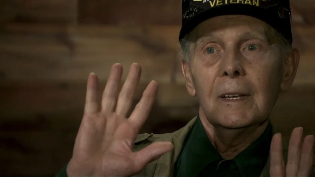 Wartorn: 1861-2010 (2010) movie still: closeup — An older man sits before a blurred wooden wall inside a dim room. He holds his hands up while…