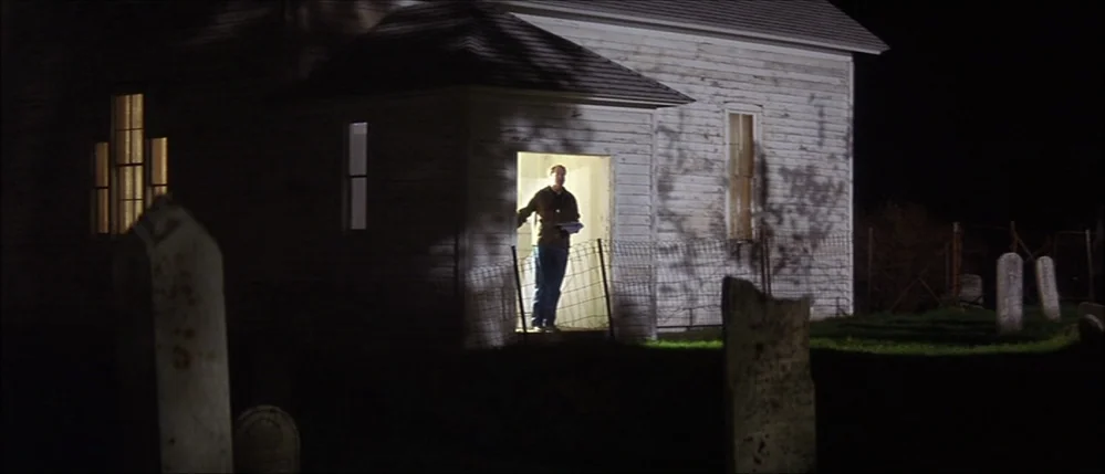 The Straight Story (1999) movie still: wide — A white wood-sided building stands in a dark cemetery at night with gravestones nearby. A lone man…