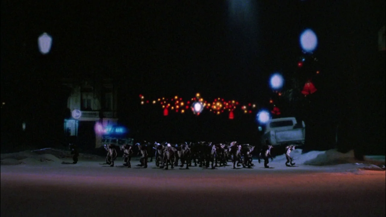 Gremlins (1984) movie still: wide — A dark, snow-covered street at night features a gathering of small monstrous creatures. Scattered holiday lights…