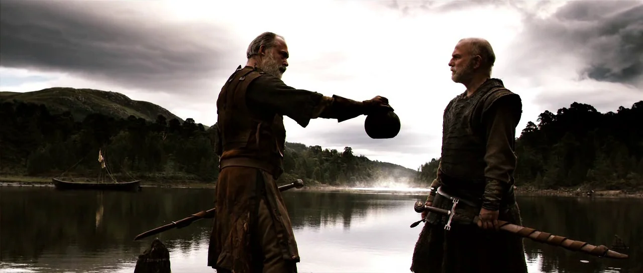 Valhalla Rising (2009) movie still: fullbody — A dark, overcast lakeside scene features two medieval men in leather armor. One man reaches out to…