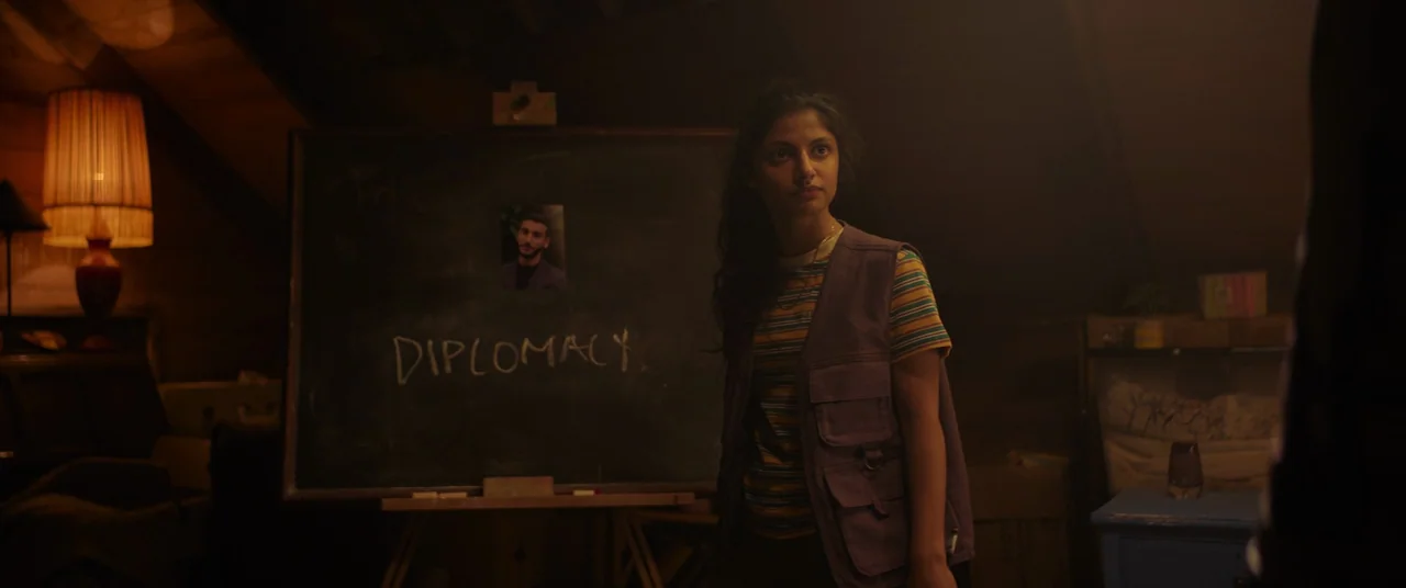 Polite Society (2023) movie still: medium — An attic room features a large chalkboard displaying a photo and the word diplomacy. A young woman…