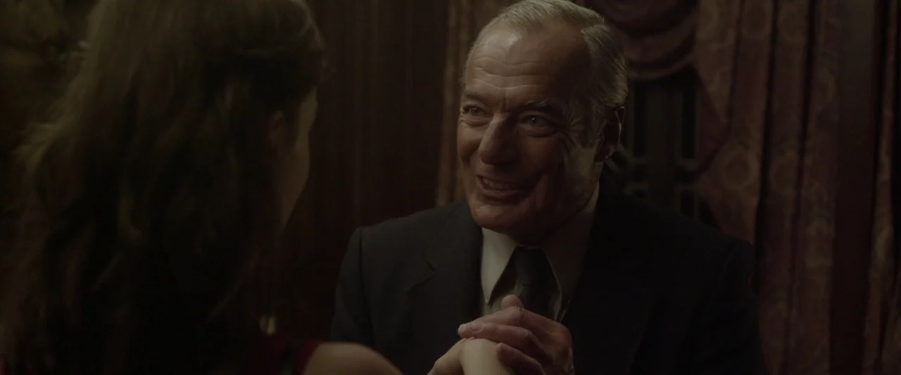 Starry Eyes (2014) movie still: medium — A dimly lit indoor space features dark, textured wall paneling and heavy fabric curtains. An elderly man in…