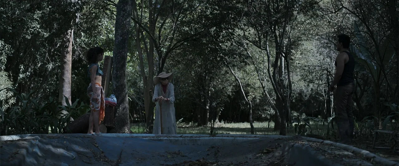 Evil Eye (2020) movie still: wide — A quiet outdoor area contains a dry concrete reservoir surrounded by dense green trees. Three people stand at…