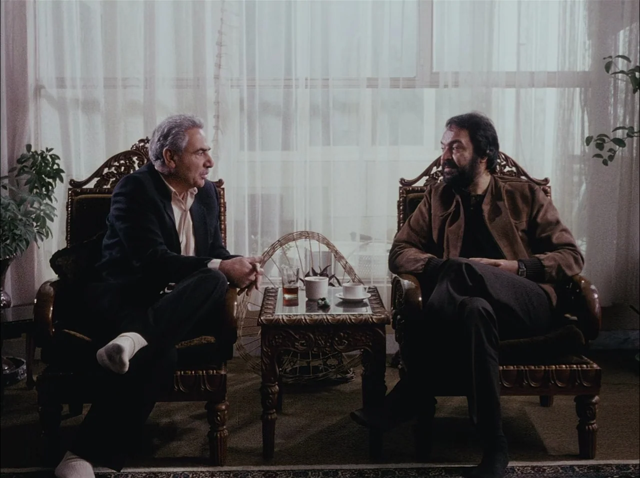 Close-Up (1990) movie still: medium — Two men sit in carved wooden armchairs with a small ornate table between them. Sheer curtains cover a large…