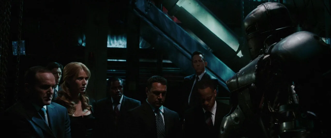 Iron Man (2008) movie still: medium — A dark industrial hangar features metal structural beams and mechanical equipment in the background. A group…