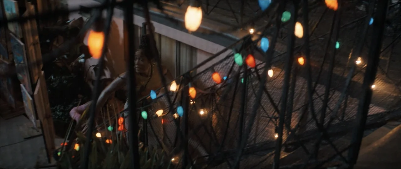 One of Them Days (2025) movie still: medium — A dark outdoor stairwell at night is decorated with various colored string lights. A young woman…