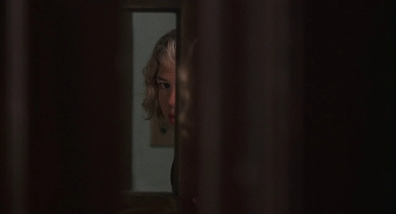 Chicago (2002) movie still: closeup — A woman looks through a thin vertical gap in a dark, wooden door. The surrounding environment is obscured by…