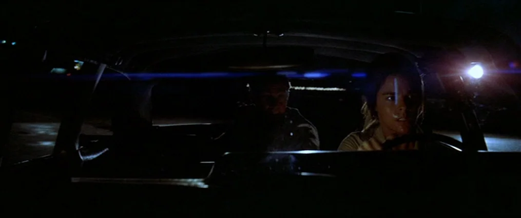 The Getaway (1972) movie still: medium — The interior of a moving car is dark, illuminated by a bright headlight ahead. A woman drives while a man…