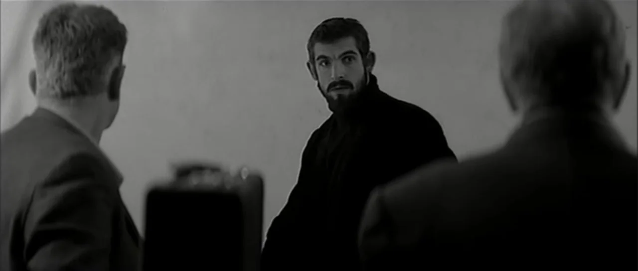 13 Tzameti (2006) movie still: medium — A stark, minimalist white interior room with no decorative details or furniture visible. A man with a beard…