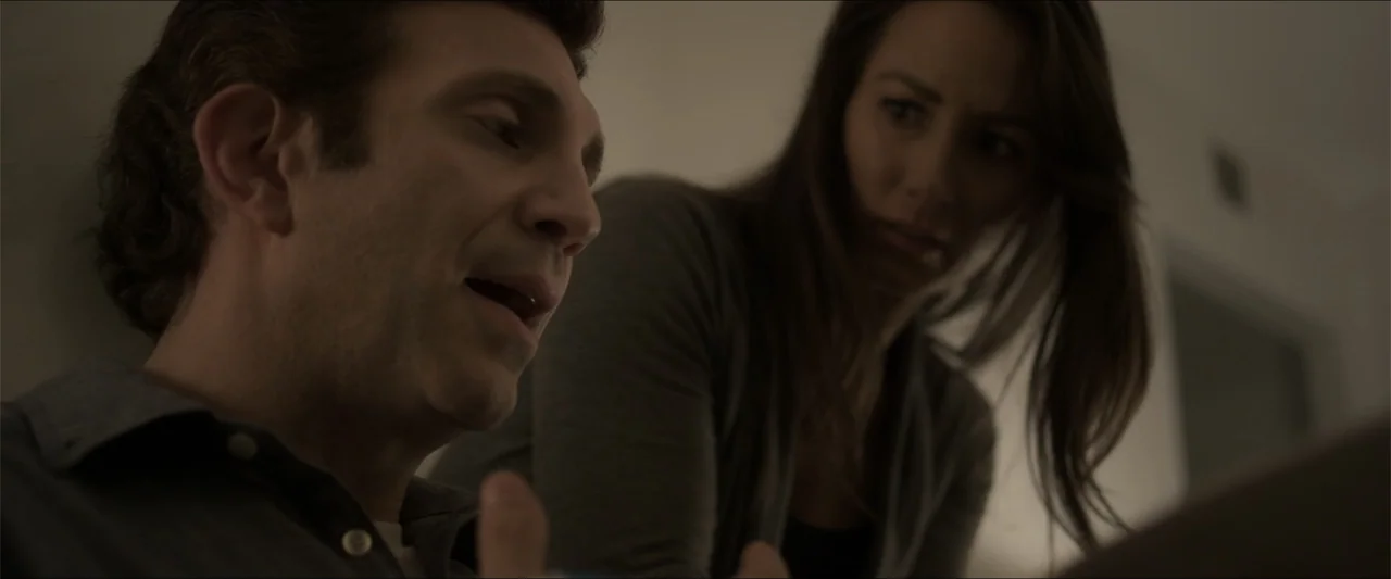 Heavenly Deposit (2019) movie still: medium — A man and a woman stand in a dimly lit interior space. The man speaks while the woman leans in and…
